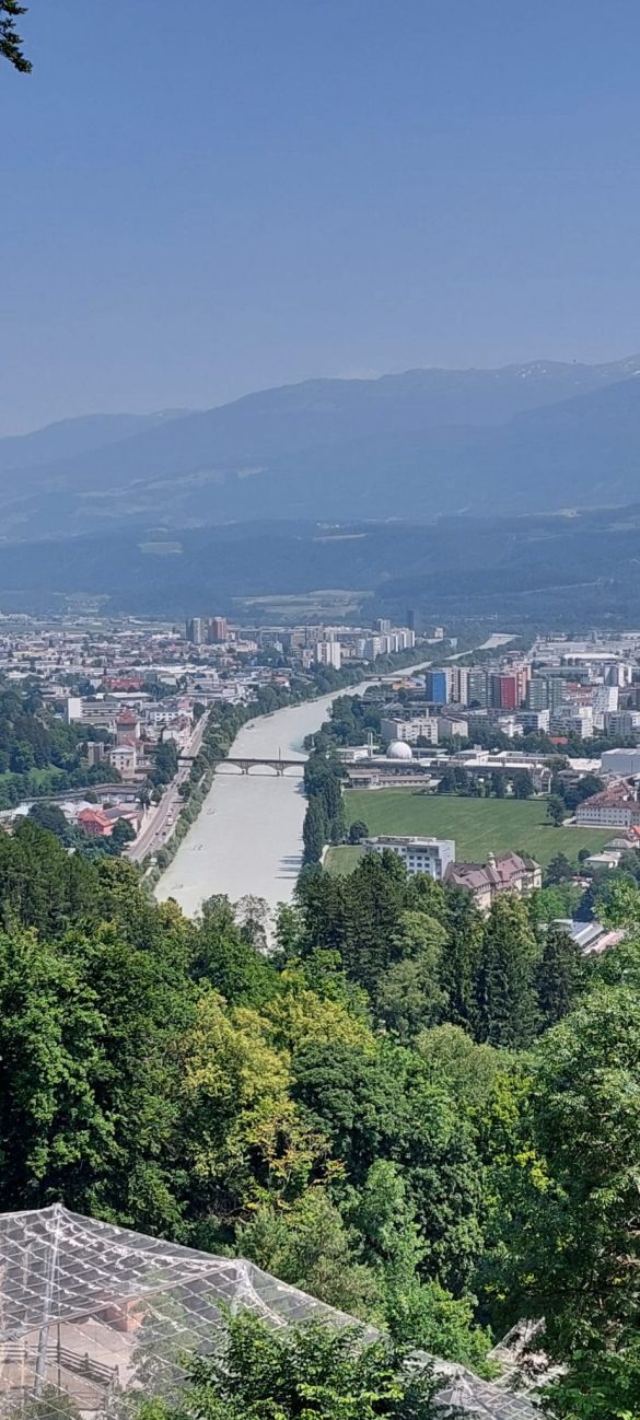 Panoramic view of Innsbruck from a high vantage point, showing the Inn River, bridges, and city buildings with mountains in the background.
